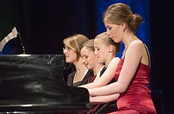 Vier Mädchen sitzen gemeinsam an einem Klavier uns spielen ein Stück vierhändig. Das Mädchen im Vordergrund trängt ein rotes glänzendes Abendkleid.