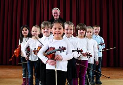 Sieben Mädchen und ein Junge stehen mit ihren Geigen V-förmig als Gruppe zusammen. Hinter ihnen steht eine Frau mit blondem Haar. Die Mädchen tragen alle weiße T-Shirts, der Junge hat ein hellblaues Hemd an.