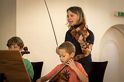 Zwei Schüler sitzen mit Violoncellos vor Notenständern. Hinter ihnen steht eine Lehrerin und spielt Geige. 